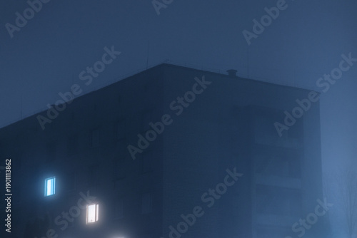 Foggy night city building with glowing windows, dark blue tones and mysterious cinematic atmosphere. Urban loneliness, isolation, insomnia, cold weather and psychological mood concept.