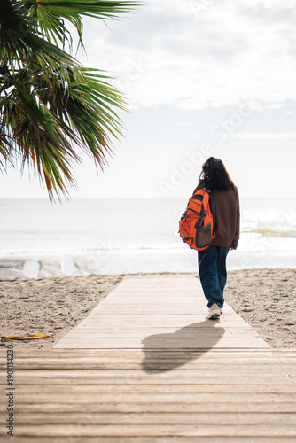 Obraz na plátně Woman with orange backpack walking along a wooden boardwalk toward the sea, rear view travel lifestyle scene with palm trees, beach, freedom, solitude and coastal vacation concept
