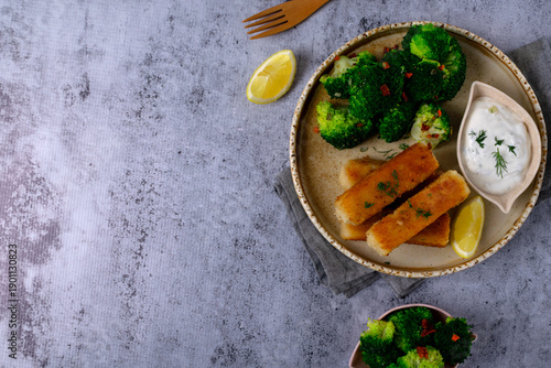 Crispy breaded fish sticks served with steamed broccoli, yogurt sauce, and a lemon wedge on a ceramic plate on a gray background. Top view, copy space