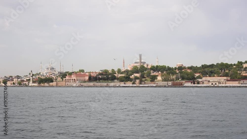 Panoramic view of Istanbul's historic district across water, featuring Hagia Sophia and Blue Mosque under cloudy sky.