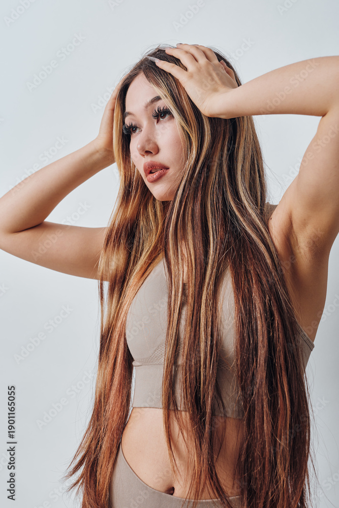 Fototapeta premium Young woman with long hair wearing beige sportswear posing with hands on head, isolated on white background for fitness or beauty concept and lifestyle portrait.