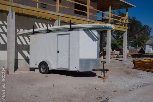 White Utility Trailer On Construction Site With Lumber And Scaffolding Under Clear Sky. Sits on a dirt lot beside a partially built structure. Lumber piles, scaffolding, and a small crane hint at ongo