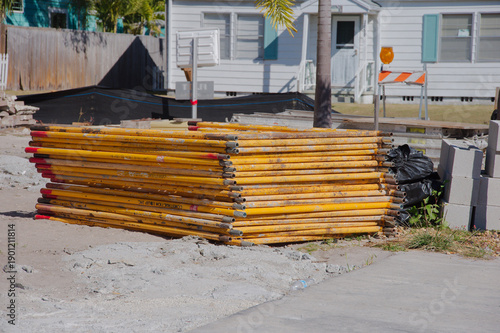 Stacked Yellow Construction Poles On A Site Near Mobile Homes And Barrier Fence.  Scaffold 
metal poles sits on a construction site by a residential area, with a palm tree, orange barrier, and stacked