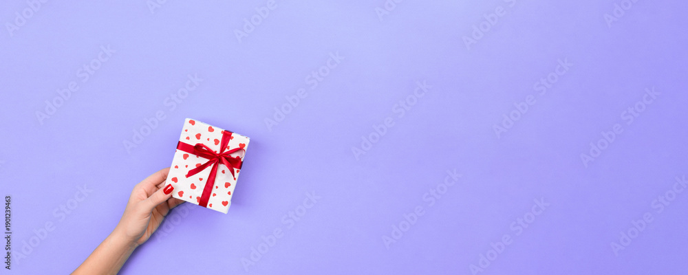 Fototapeta premium Woman hands give wrapped valentine or other holiday handmade present in paper with red ribbon. Present box, red heart decoration of gift on violet table, top view with copy space for you design