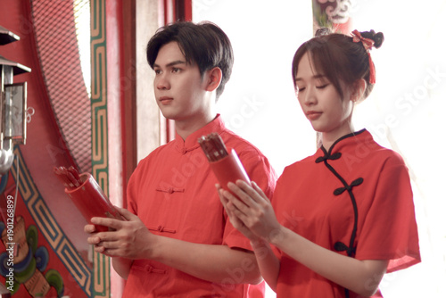Happy Asian couple in traditional Chinese red dress paying respects at shrine for good fortune in celebrating the Chinese New Year. Lovers casting Chi Chi fortune sticks during make wishes together.