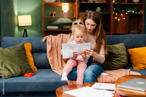 Mother and daughter reading paper document together while relaxing on sofa at home