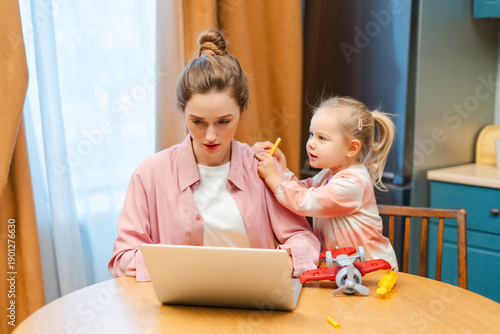 Mother working on laptop while daughter seeking attention, balancing work and family