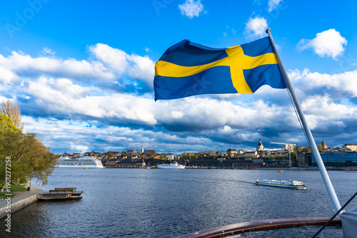 Swedish flag waving in the wind against a backdrop of dark, dramatic cumulus clouds at the stern of the ship. In the background is the Stockholm district of Slussen. Sweden, pnational pride