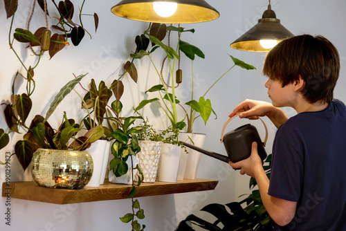 Teenage boy watering indoor plants at home, sustainable lifestyle and indoor gardening