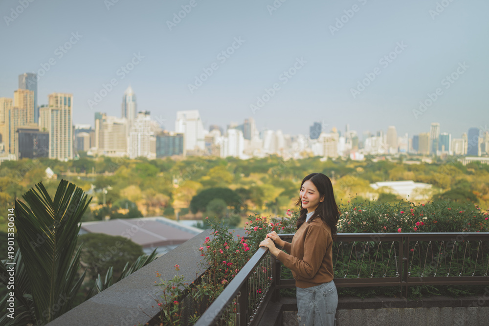 Naklejka premium Happy Asian woman standing on rooftop terrace overlooking city skyline and public park. Urban lifestyle relaxation and travel concept. Female enjoying panoramic view of skyscrapers and green nature