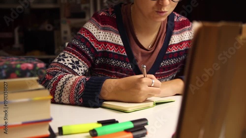 Young woman in glasses and a cozy patterned sweater writing in a notebook. Studying or journaling at home