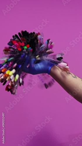 Hand in a blue glove holding a large fan of colorful nail polish samples against a pink background. Manicure concept. Vertical video
