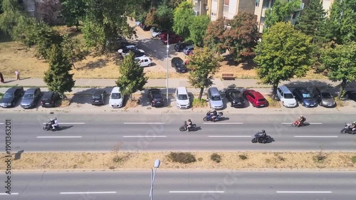 A top-down, high-angle shot captures a group of bikers riding along a multi-lane urban avenue. Closely following behind the motorcycles is a police patrol vehicle. 