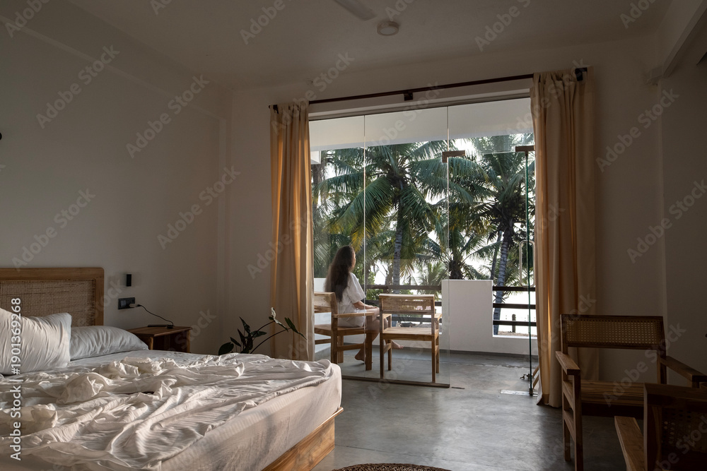 Fototapeta premium A young woman with long dark hair and a white T-shirt walks out onto the balcony from her luxurious bed after waking up in her hotel room. Wake-up time in a hotel room in Sri Lanka on vacation.