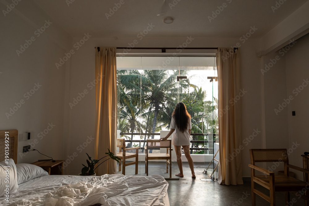 Fototapeta premium A young woman with long dark hair and a white T-shirt walks out onto the balcony from her luxurious bed after waking up in her hotel room. Wake-up time in a hotel room in Sri Lanka on vacation.