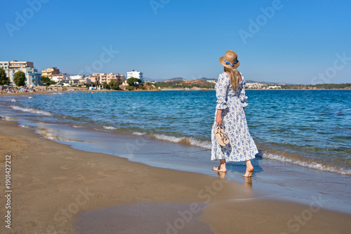 Fotografía Woman in a light summer dress and straw hat walking barefoot near the shoreline on a Greek beach