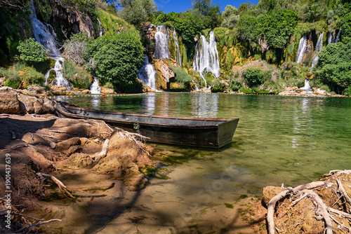 Scenic Kravica waterfalls with cascading streams, green lagoon, and wooden boat in natural surroundings.