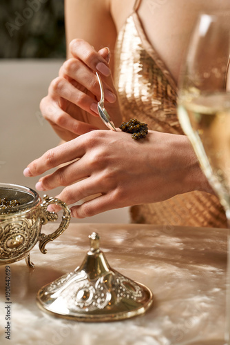 Luxury lifestyle: Woman in a gold sequin dress placing black sturgeon caviar on her hand with a silver spoon.