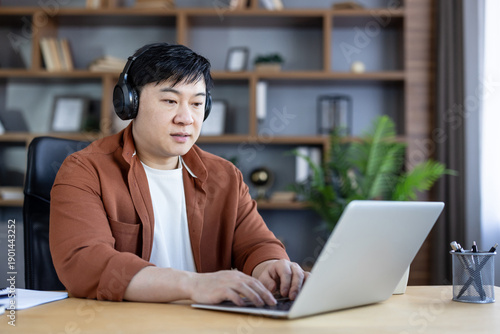 Fotografie Asian man with headphones concentrating on his laptop, typing and working remote