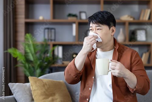 Asian man feeling unwell on a sofa, blowing his nose into a tissue while holding a warm mug, resting at home with cold or flu symptoms, seeking comfort and recovery