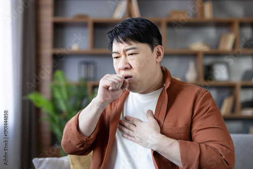 Schilderij op canvas Asian man at home clutching chest and covering his mouth while coughing, showing