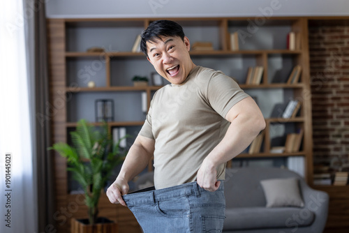 Fotografija Asian man happily showing significant weight loss results by holding out his old