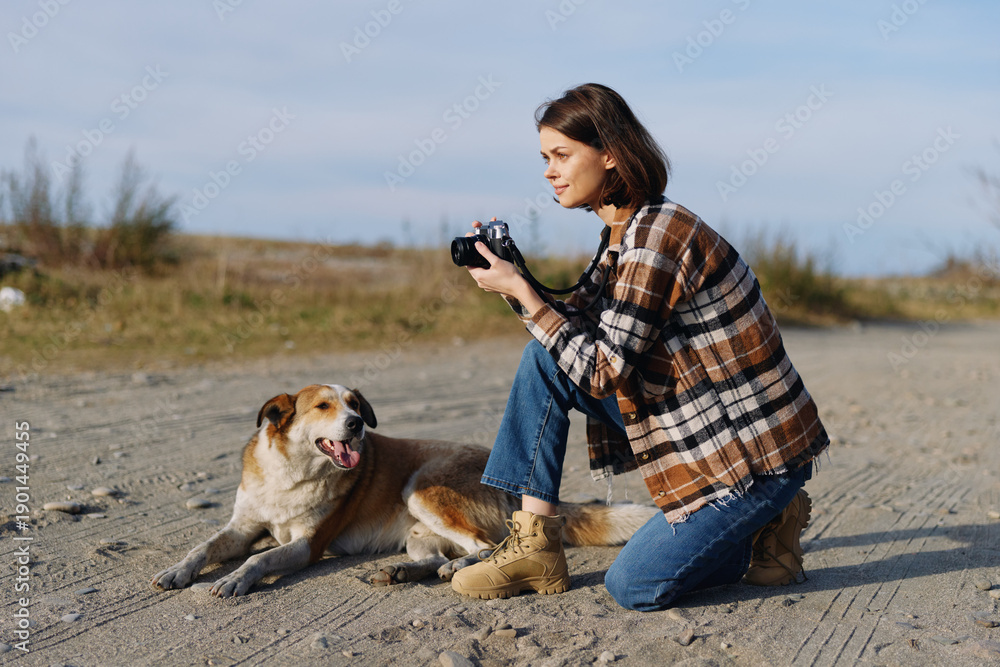 Fototapeta premium woman photographer kneels on sandy outdoor terrain with a dog beside her holding a camera and focusing on subjects in the open landscape under a clear sky