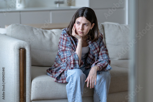 Canvastavla Thoughtful bored young woman sitting on sofa at home, resting face on hand with distant look