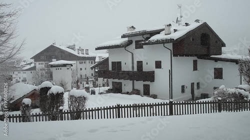 Viseo of snow-covered alpine houses sit quietly during a winter storm, viewed through window with curtains. Soft snowfall, muted light, and wooden chalets create a calm, cold, and secluded mountain