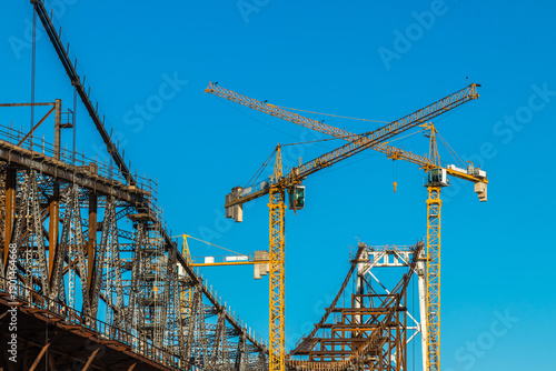 Photos Steel bridge construction with towering cranes against clear blue sky, showcasin