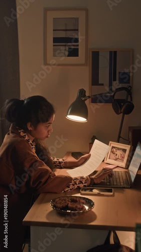 Vertical shot of young adult multiethnic woman typing on laptop keyboard entering data while analyzing document, remote employee working at cozy desk with lamp in home office