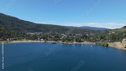Aerial view of Lake Lacar and San Martin de los Andes in Patagonia, highlighting a scenic marina with numerous sailboats and boats anchored in deep blue water surrounded by mountains.