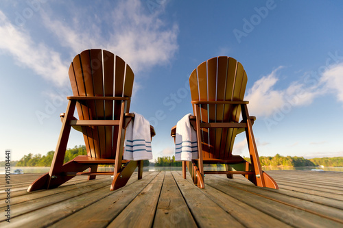 Low-angle view of a weathered dock with two Adirondack chairs and striped towels. Wooden planks lead to a calm lake and lush green shoreline under a bright summer sky. A peaceful, sun-drenched morning