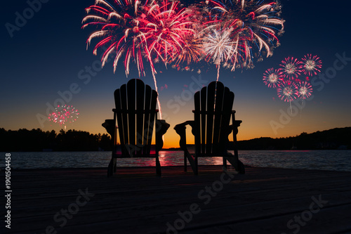 Night scene in Muskoka with two Adirondack chairs silhouetted on a dock. Fireworks burst over a calm lake, celebrating Canada Day at a summer cottage. Ideal for festive and seasonal holiday themes.