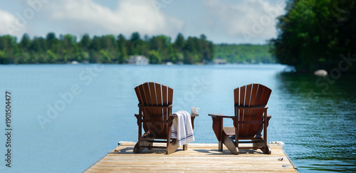 Wide-angle view of a wooden dock with two Adirondack chairs facing a calm lake. A striped towel suggests a relaxing morning. Lush forest and bright sky create a peaceful atmosphere with copy space.