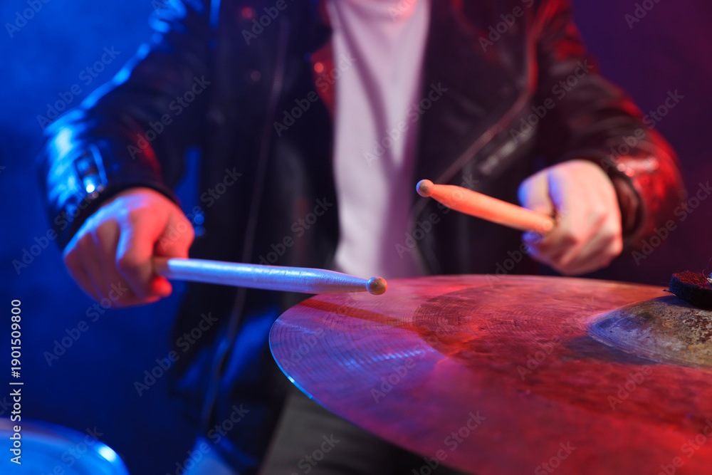 Obraz premium Man with drumsticks playing cymbal in color lights against dark background, closeup