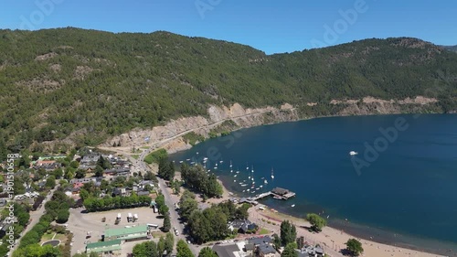 Aerial view of Lake Lacar and San Martin de los Andes in Patagonia, highlighting a scenic marina with numerous sailboats and boats anchored in deep blue water surrounded by mountains.