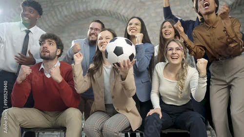 A medium group of young, diverse office colleagues watching a football match, cheering, and one person holding a football. Energetic workplace scene showing team spirit, fun.