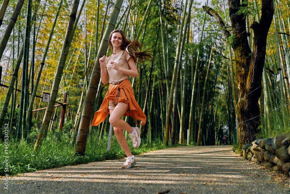 Fototapeta premium Woman running in bamboo forest path, smiling and wearing orange skirt and sneakers, outdoor jog for fitness and travel, healthy young adult enjoying nature and sunlight on gravel trail