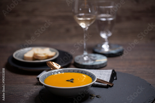 Kürbissuppe in einer Suppenschale auf einem gedeckten Tisch vor einem Hintergrund aus Holz. Nahaufnahme der Suppe, zwei Gäser und ein Schälchen mit Brot, Tischsituation. 