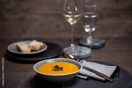 Kürbissuppe in einer Suppenschale auf einem gedeckten Tisch vor einem Hintergrund aus Holz. Nahaufnahme der Suppe, zwei Gäser und ein Schälchen mit Brot, Tischsituation. 