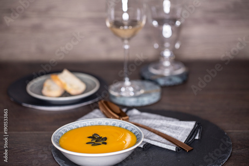 Kürbissuppe in einer Suppenschale auf einem gedeckten Tisch vor einem Hintergrund aus Holz. Nahaufnahme der Suppe, zwei Gäser und ein Schälchen mit Brot, Tischsituation. 