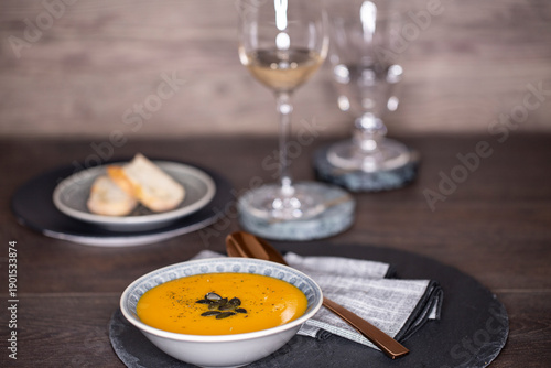 Kürbissuppe in einer Suppenschale auf einem gedeckten Tisch vor einem Hintergrund aus Holz. Nahaufnahme der Suppe, zwei Gäser und ein Schälchen mit Brot, Tischsituation. 