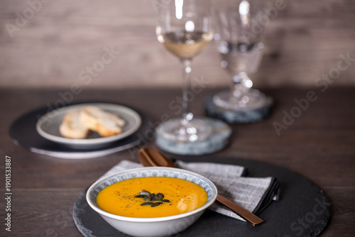 Kürbissuppe in einer Suppenschale auf einem gedeckten Tisch vor einem Hintergrund aus Holz. Nahaufnahme der Suppe, zwei Gäser und ein Schälchen mit Brot, Tischsituation. 