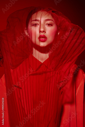 Fashion model in extravagant red Costume poses behind a sheer red fabric, emphasizing bold makeup and avant-garde style. Creative beauty and fashion editorial concept in studio lighting.
