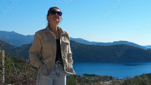 Tourist enjoying breathtaking lake view. Woman traveler standing near mountain lake, wearing sunglasses and enjoying scenic landscape on bright summer day, embodying peaceful wanderlust