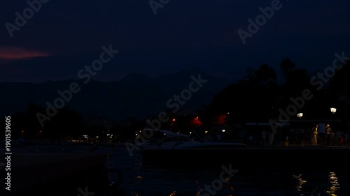 People walking along an illuminated night pier. Night view of a calm harbor with people strolling along the illuminated pier, with distant mountains silhouetted against the dark blue twilight sky