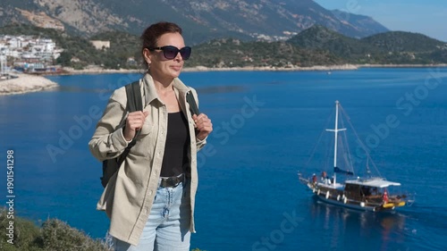 Tourist enjoying breathtaking seascape with anchored boat. Female traveler wearing sunglasses standing on viewpoint, enjoying panoramic seascape with traditional wooden turkish gulet