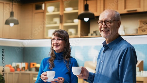 Portrait of senior joyous couple enjoying retired life in the kitchen, smiling by the counter and serving coffee. Mature people embrace relaxed moments for joyful lifestyle and wellbeing. Camera B.