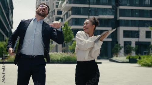 Two joyful active multiracial business colleagues dancing outside city celebrating success Caucasian businessman African businesswoman coworkers male female dance victory party corporate fun movement